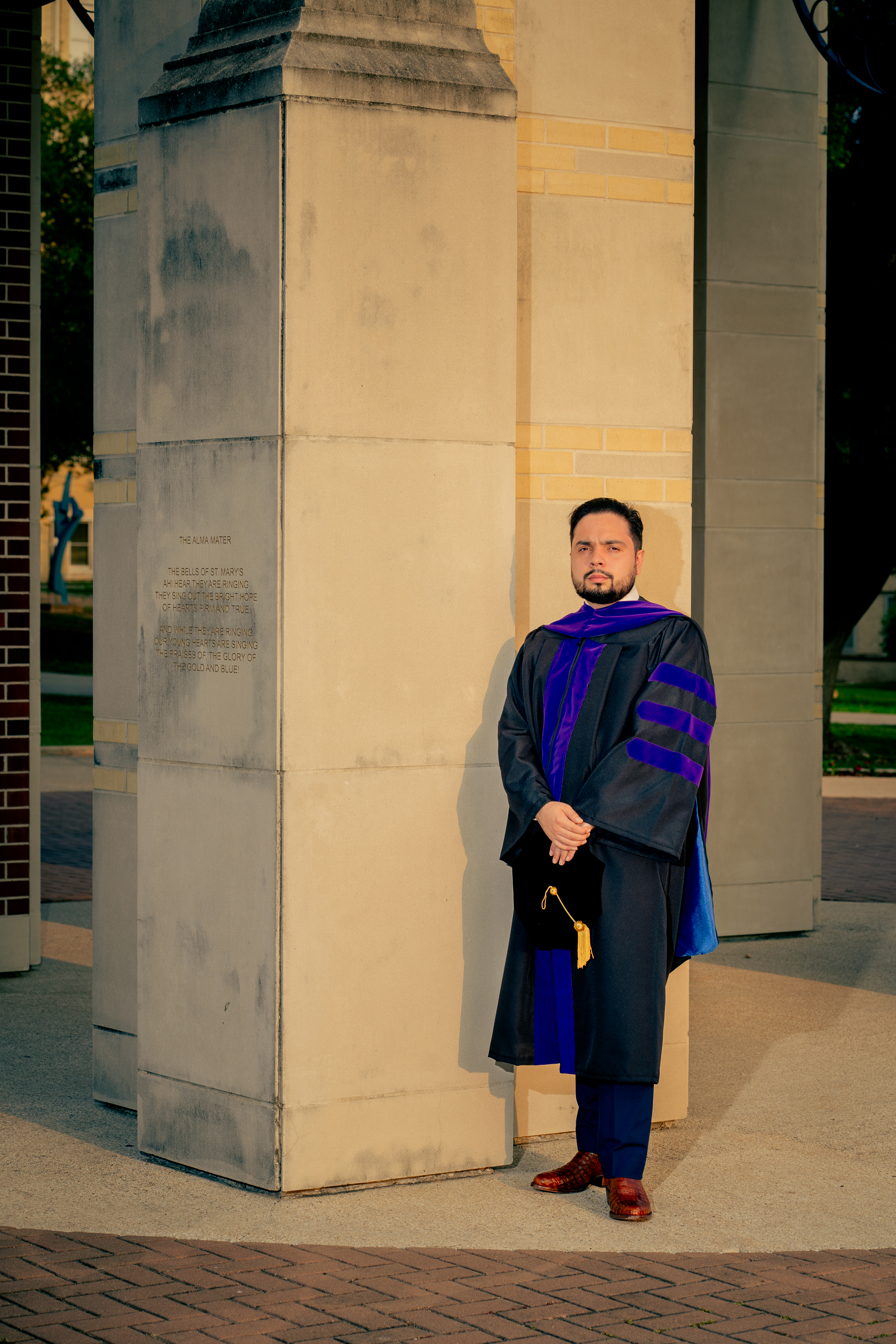 Doctoral graduate standing beside the Alma Mater pillar in cap and gown, St. Mary's University