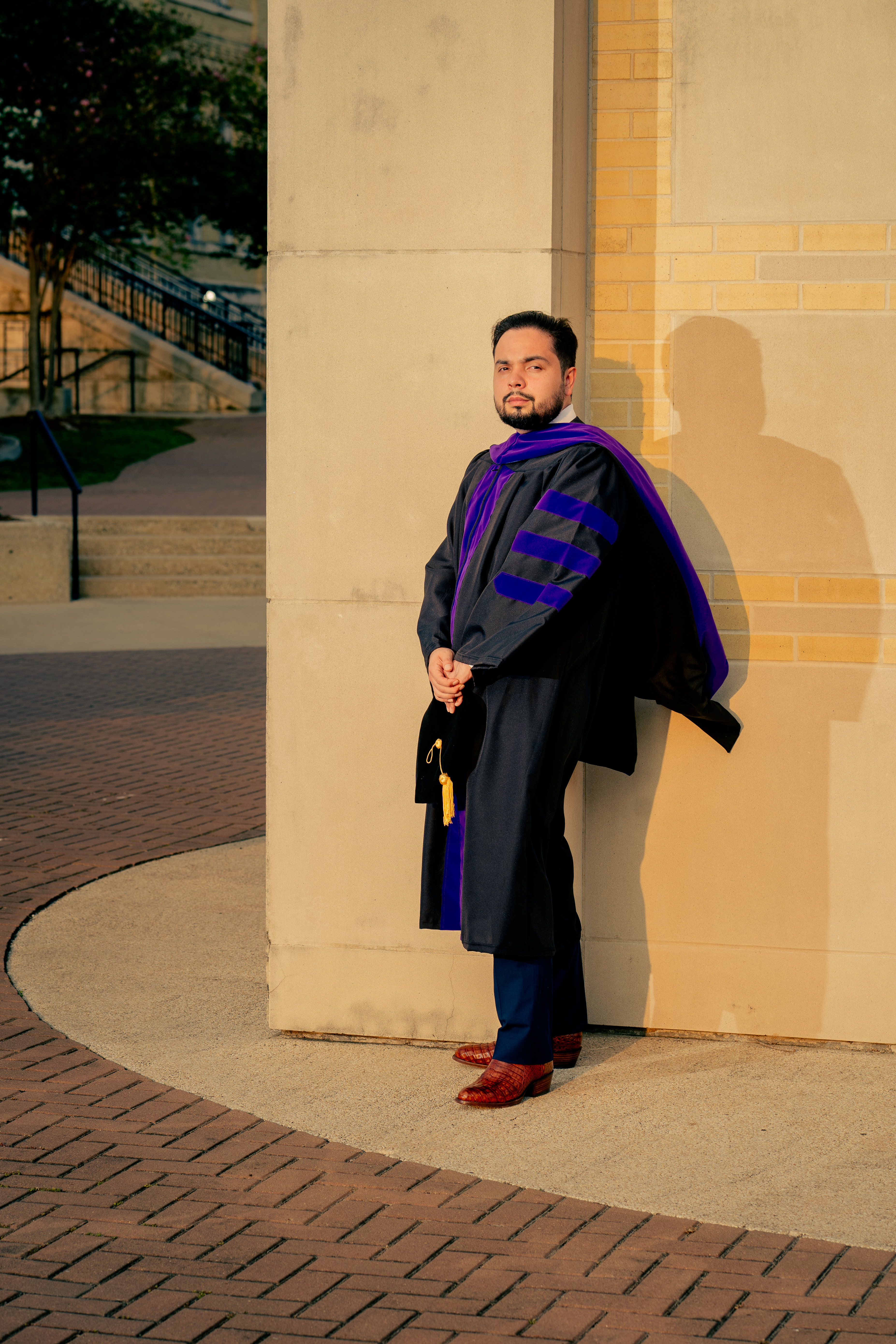 Graduate in purple-and-black doctoral hood leaning against a campus column at golden hour