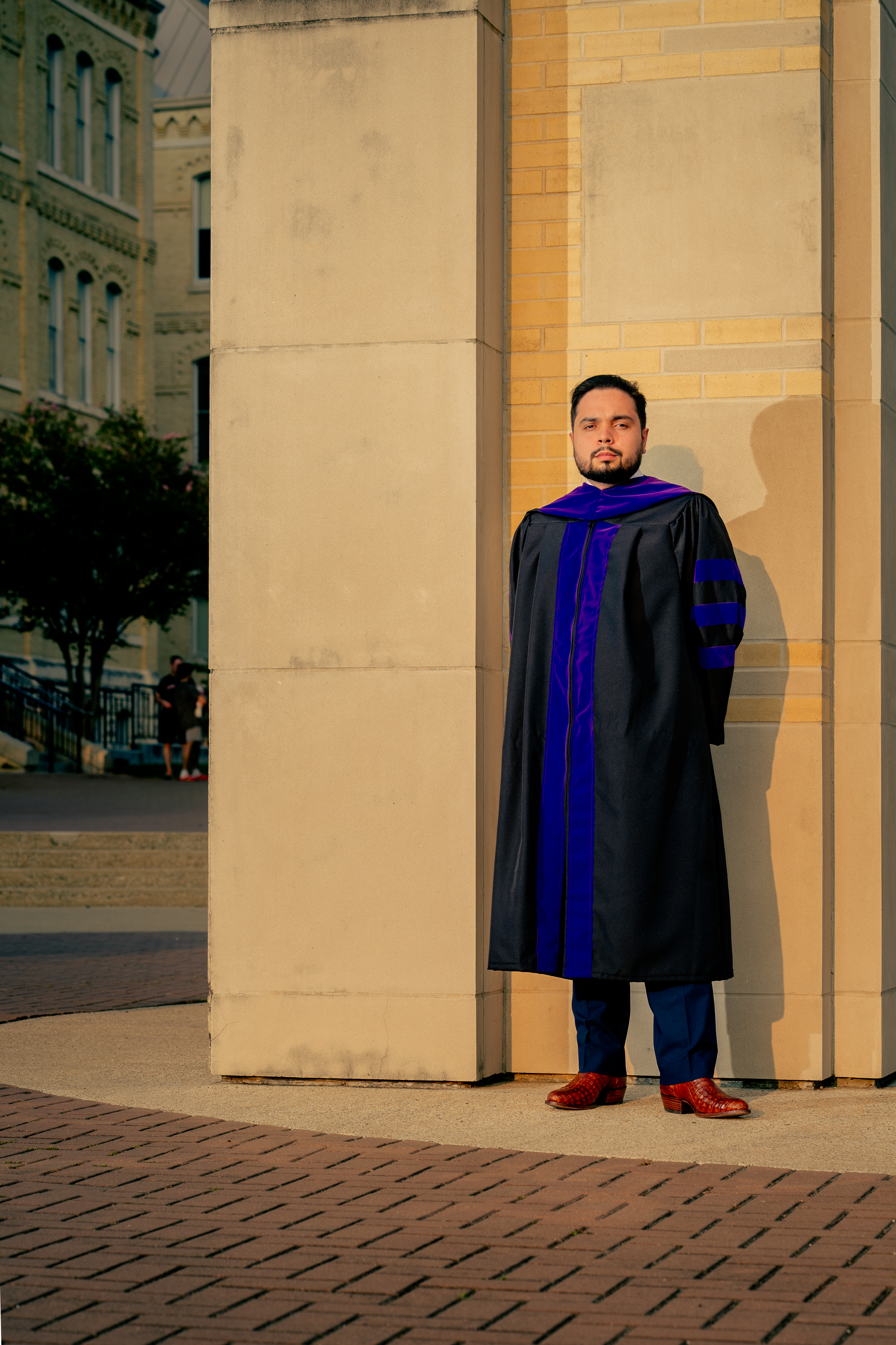Front-facing portrait of doctoral graduate in full regalia against a sunlit campus wall