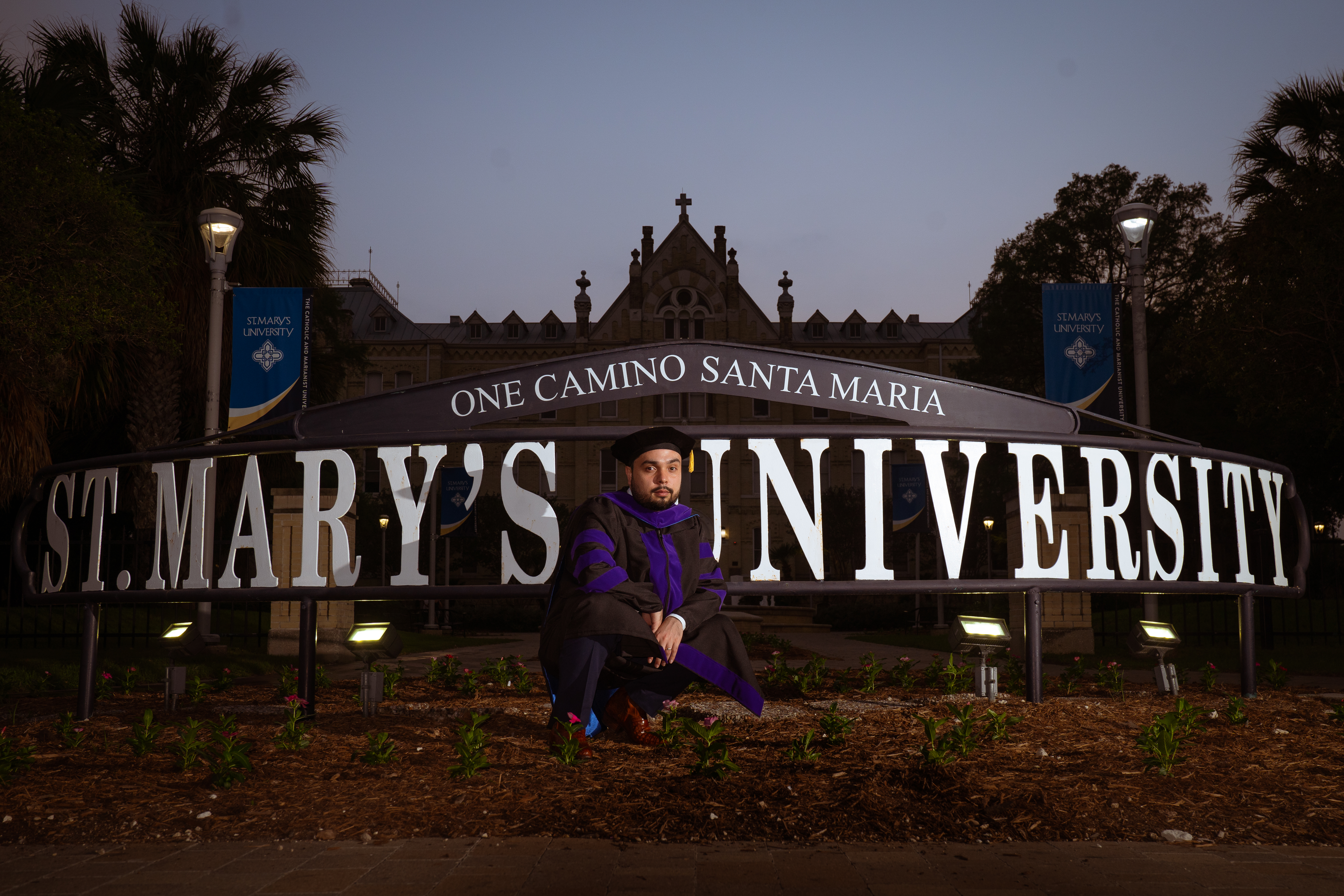 Graduate in doctoral regalia kneeling in front of the illuminated St. Mary's University sign at dusk, San Antonio TX