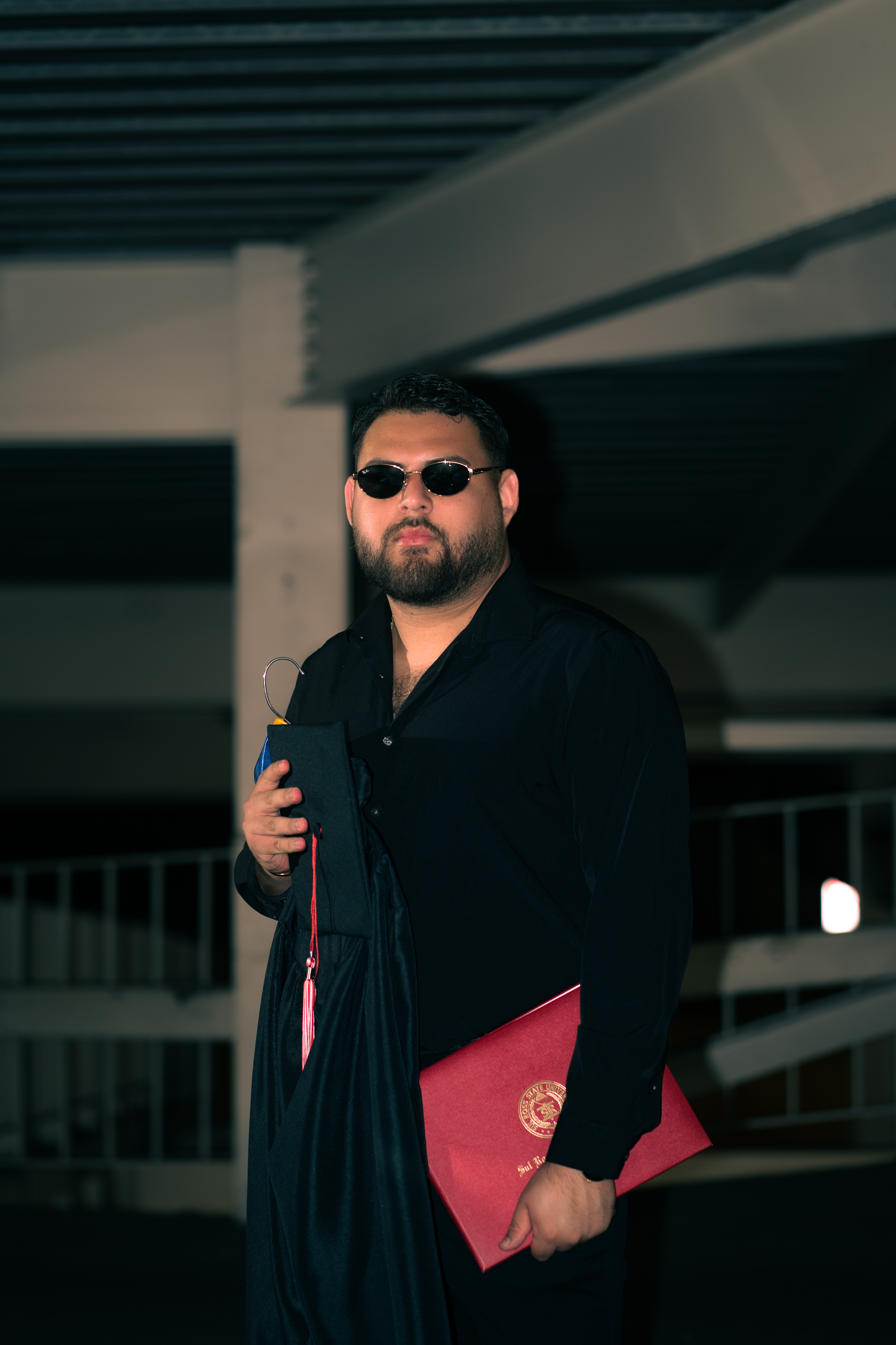 Graduate in black shirt and sunglasses holding cap and red diploma folder in a parking garage at night, Texas State University