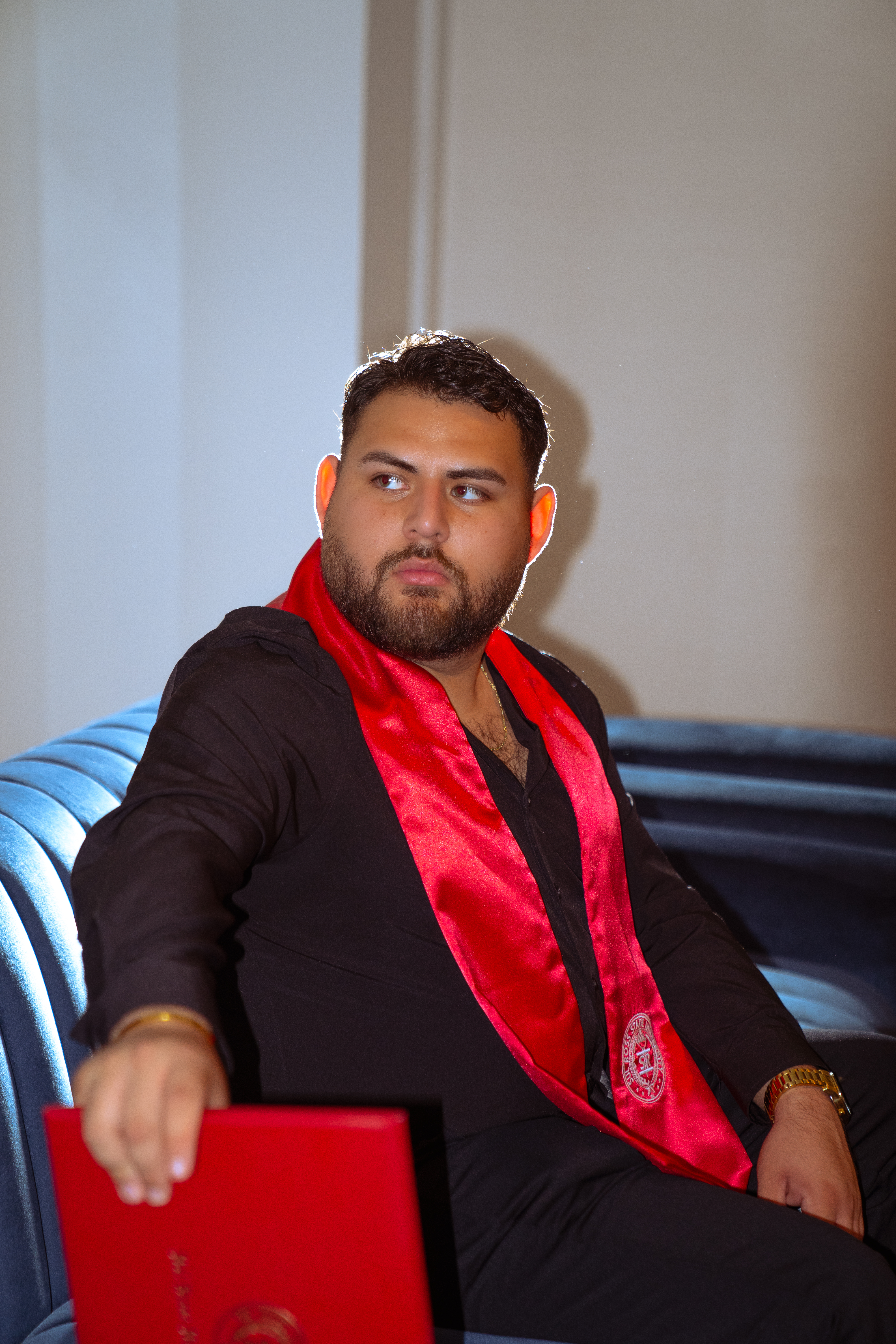 Graduate seated on a blue velvet sofa wearing a red Texas State University stole, holding a diploma folder