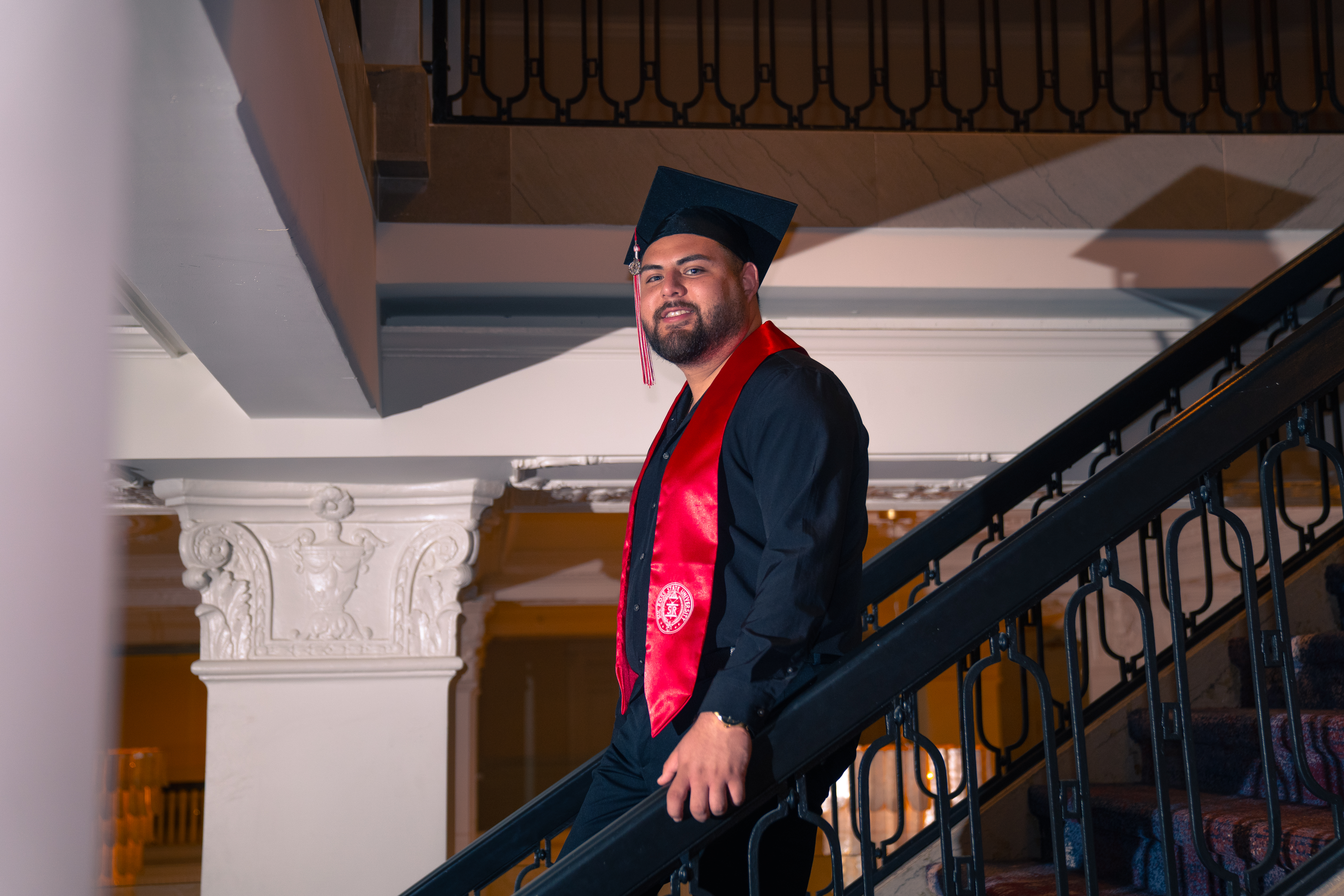 Graduate seated, adjusting his graduation cap with red tassel against a sunlit wall