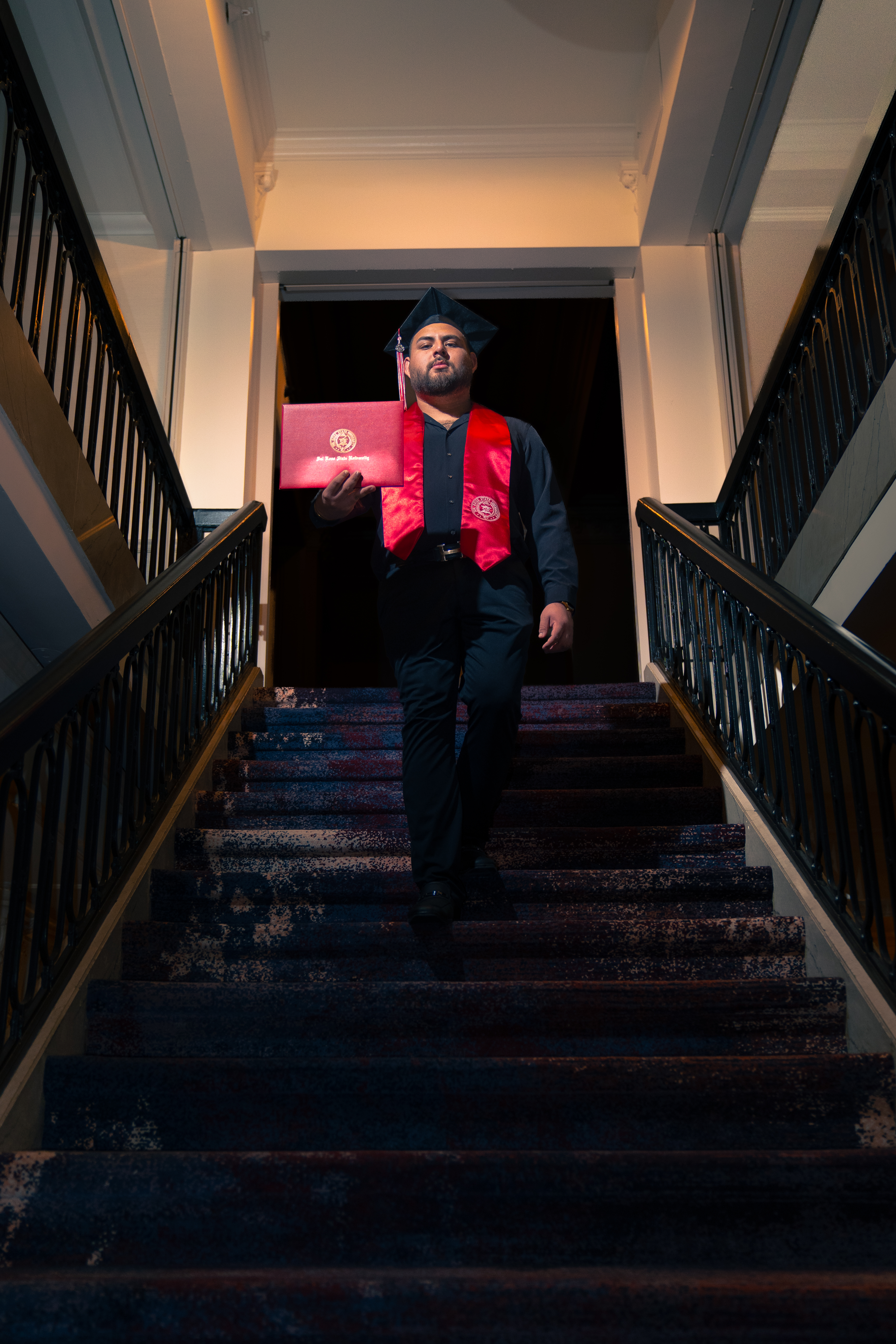 Graduate in cap and red stole descending a patterned carpeted staircase holding a red diploma