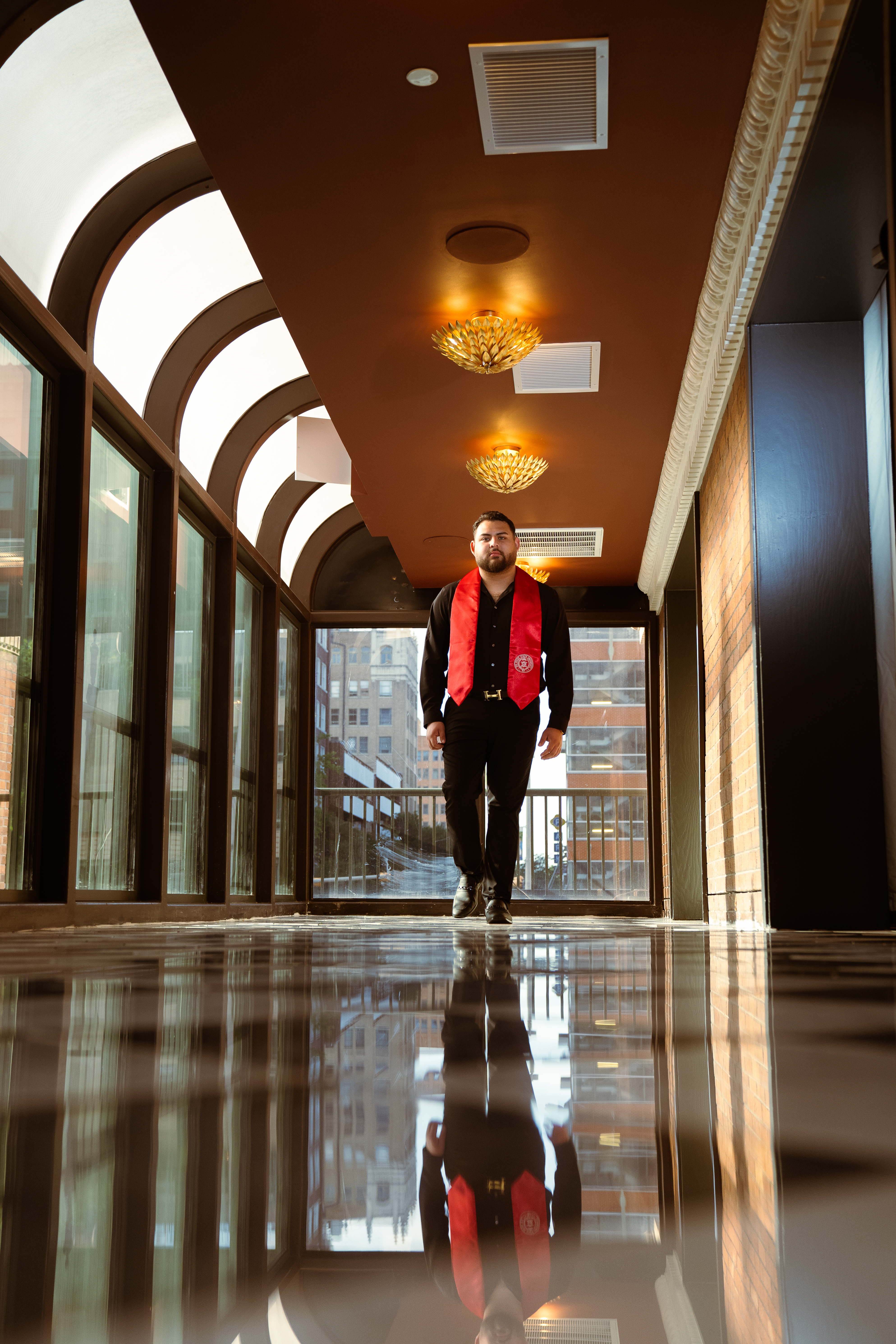 Graduate in red stole walking down an arched, sunlit skywalk with reflective floor, downtown San Antonio
