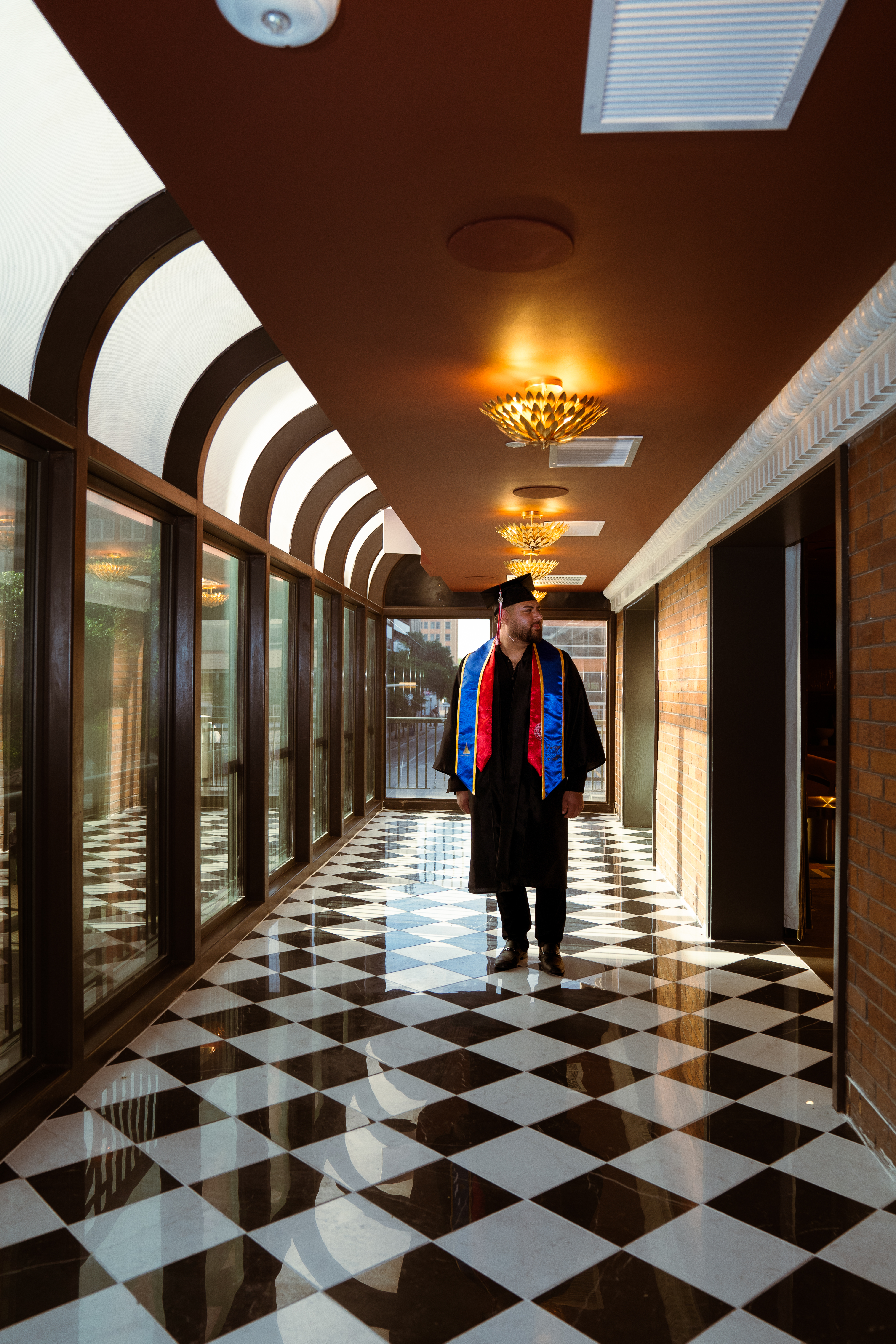 Graduate in full gown and honor stoles standing on a black-and-white checkered marble floor in an arched corridor
