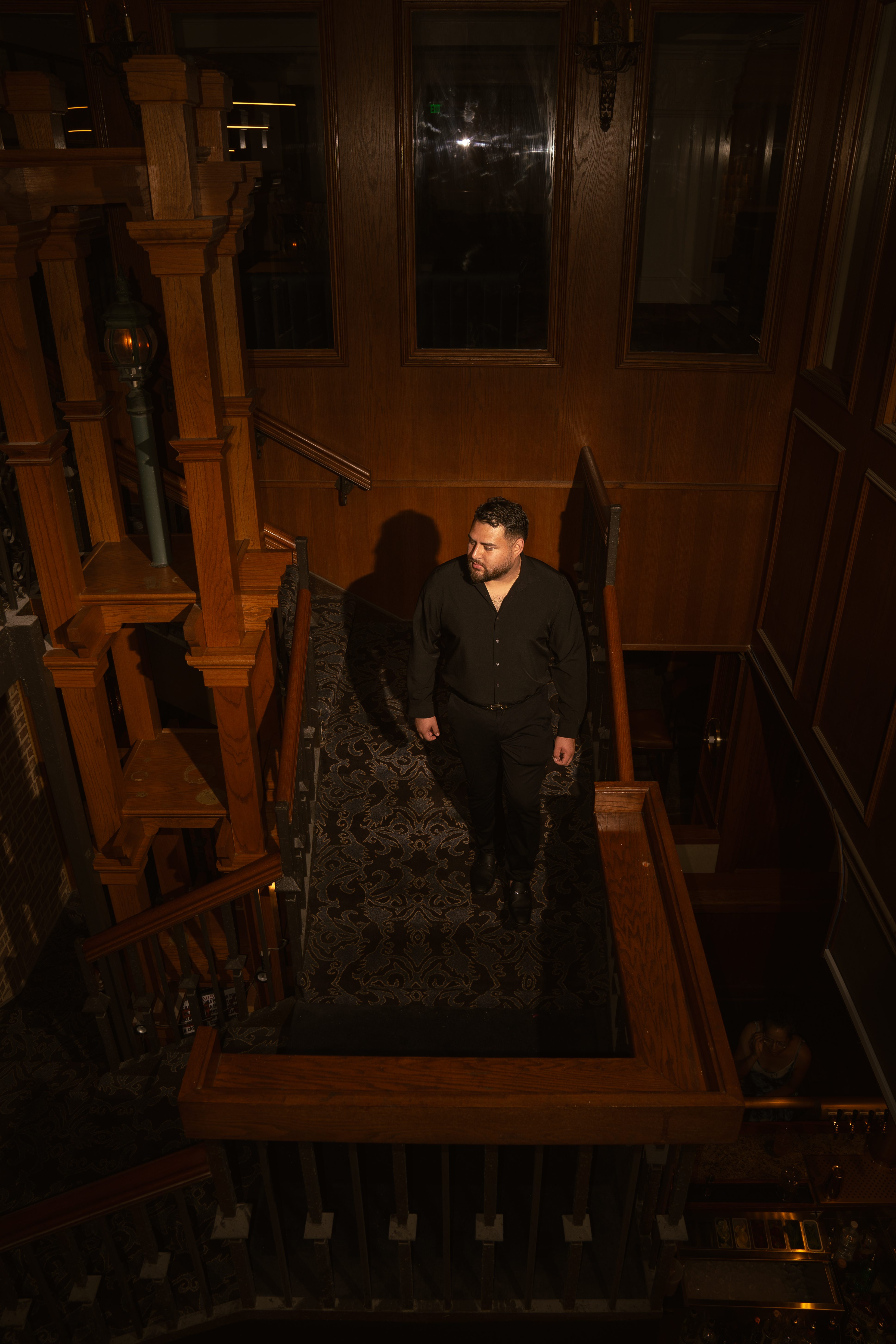 Graduate in black descending a moody wood-paneled staircase under a single spotlight