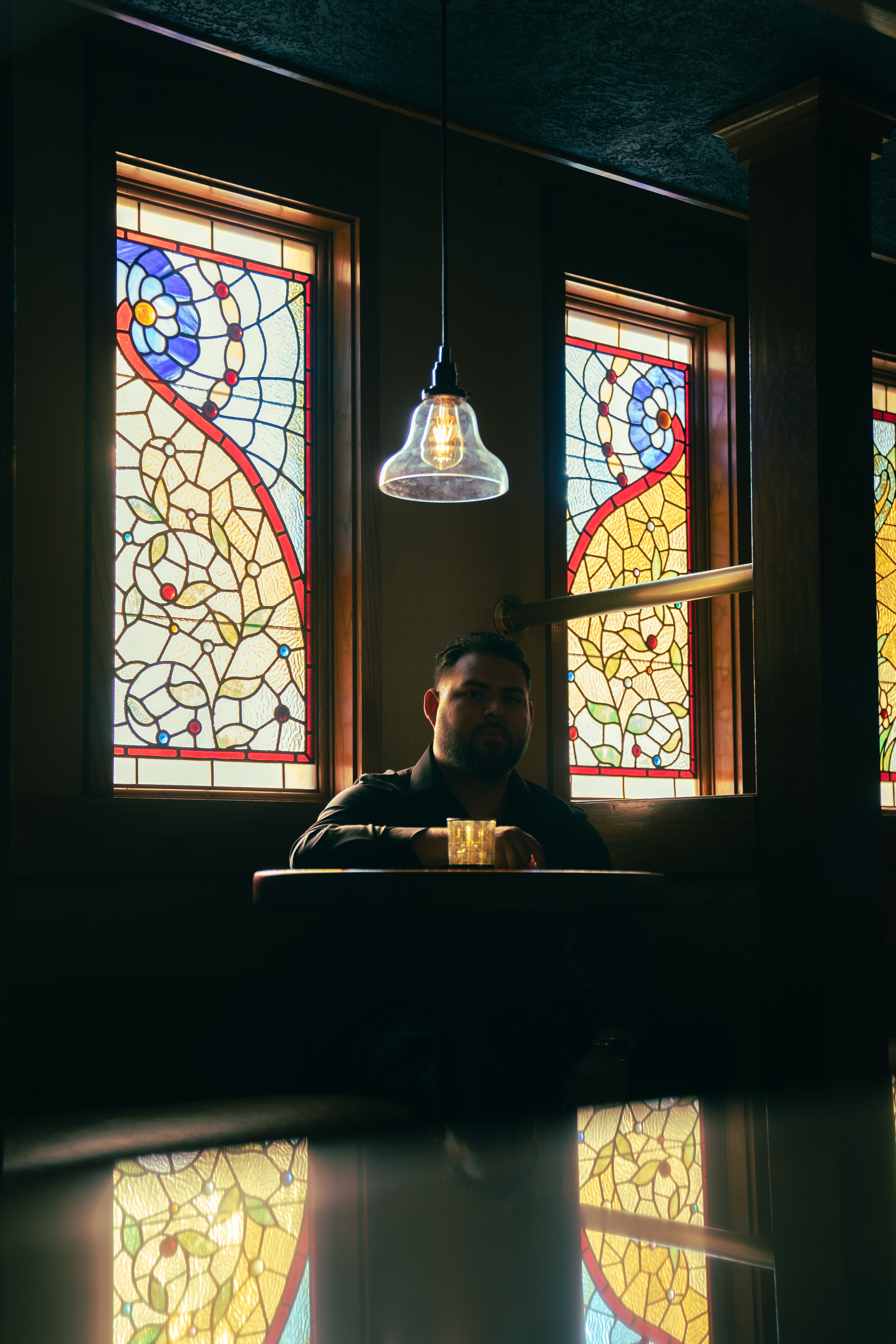 Graduate seated at a bar table framed by ornate stained-glass windows holding a glass